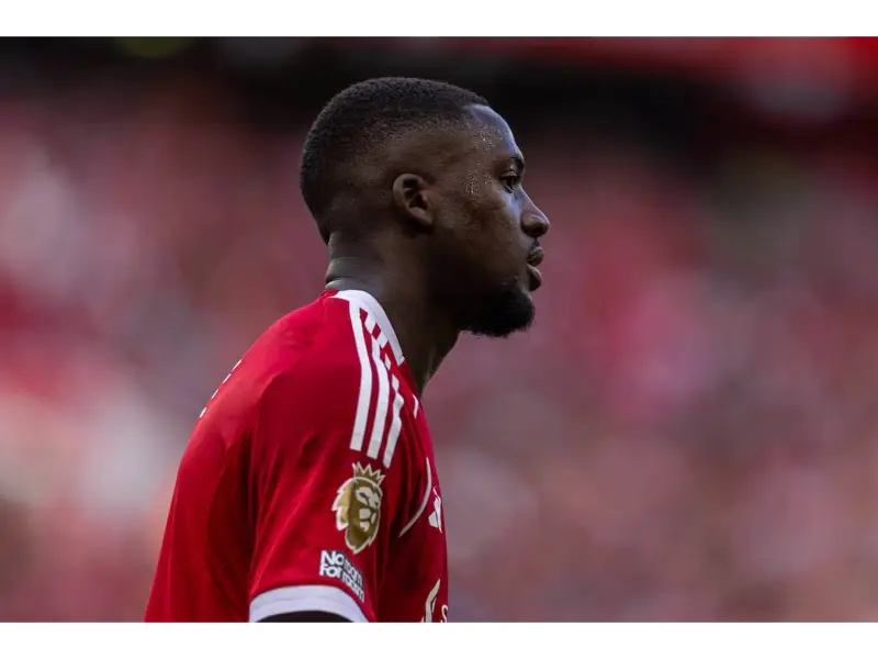Side profile of a Liverpool defender in a red home kit, sweating and looking focused during a Premier League match with a blurred red crowd in the background.