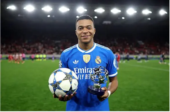 Real Madrid player Mbappé in blue away kit holding the match ball in one hand and a Champions League Player of the Match  Olympiacos vs Real Madrid analysis trophy in the other on the pitch.