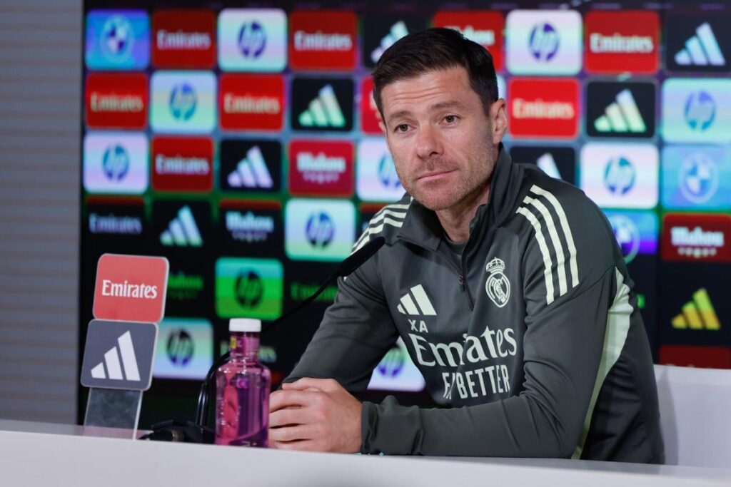 Coach in a Real Madrid tracksuit speaks at a press conference podium with sponsor backboard.