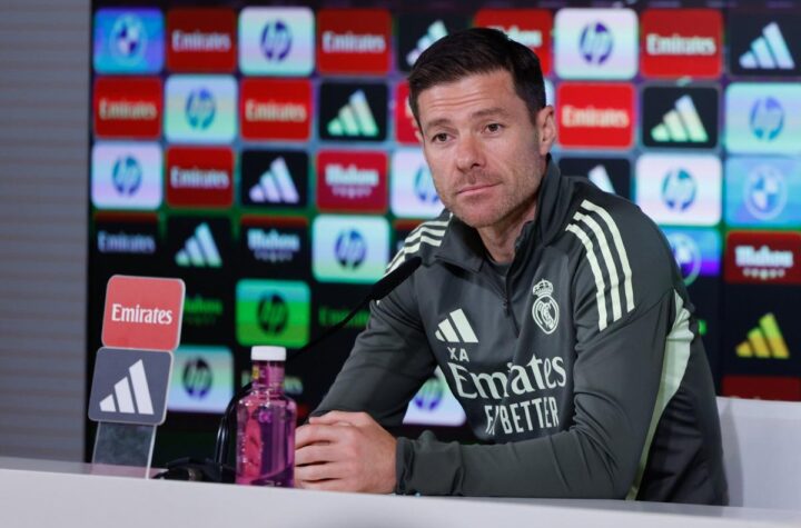 Coach in a Real Madrid tracksuit speaks at a press conference podium with sponsor backboard.
