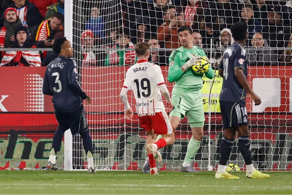 Real Madrid goalkeeper catches the ball inside the box while a Girona forward and two Madrid defenders look on during a tense La Liga match in Girona.