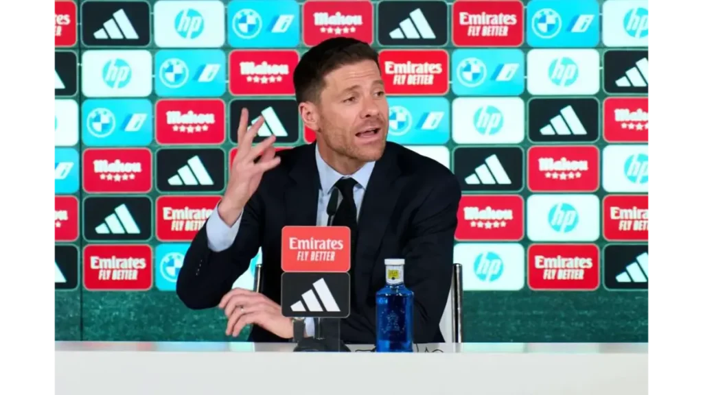Xabi Alonso gesturing while speaking at a Real Madrid press conference in front of sponsor backdrop