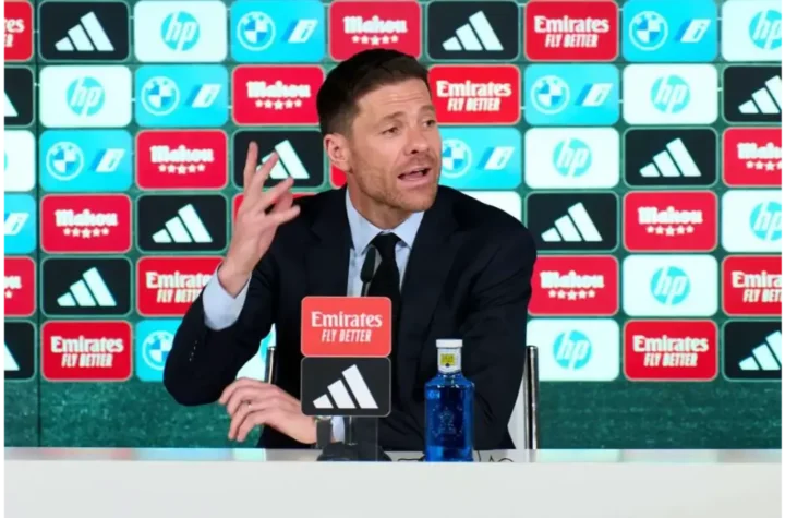 Xabi Alonso gesturing while speaking at a Real Madrid press conference in front of sponsor backdrop