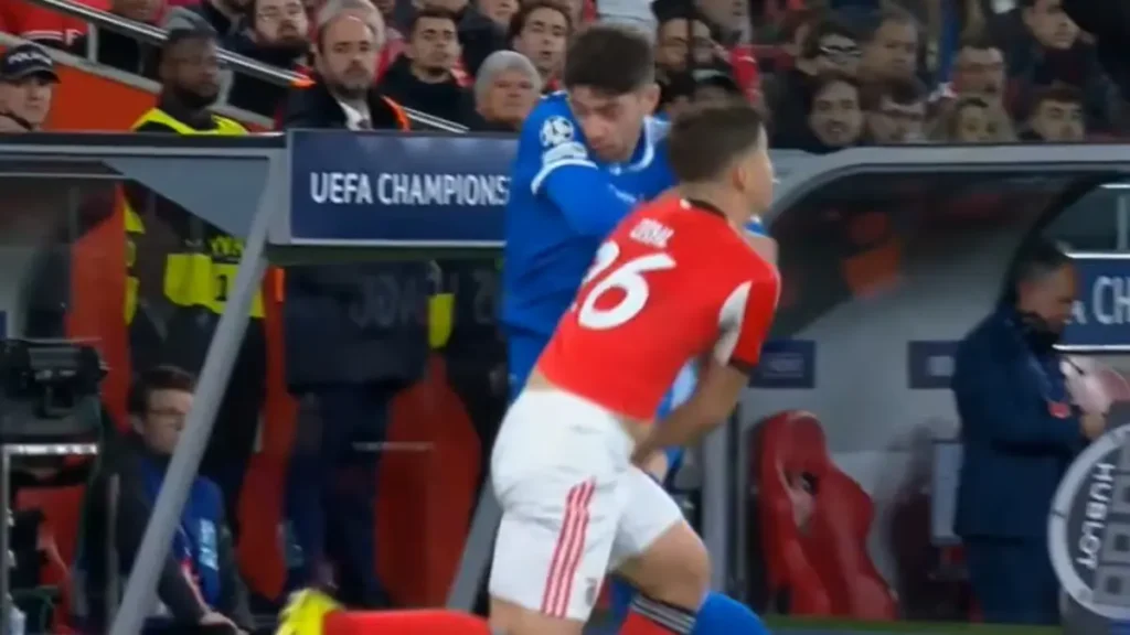 Red-shirted player and blue-shirted opponent collide near the sideline during a UEFA Champions League match, with the bench area visible in the background.