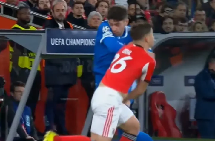 Red-shirted player and blue-shirted opponent collide near the sideline during a UEFA Champions League match, with the bench area visible in the background.