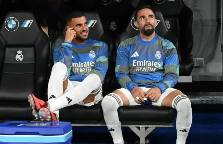 Two Real Madrid players sit on the bench before kickoff wearing blue training tops and white socks