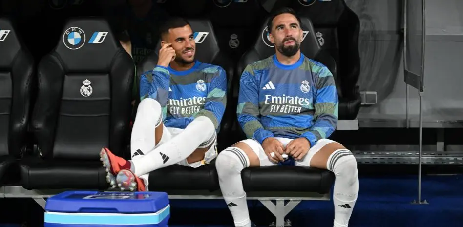 Two Real Madrid players sit on the bench before kickoff wearing blue training tops and white socks