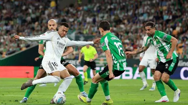 A Real Madrid player in white shoots the ball while surrounded by several Real Betis defenders in green during a La Liga match.