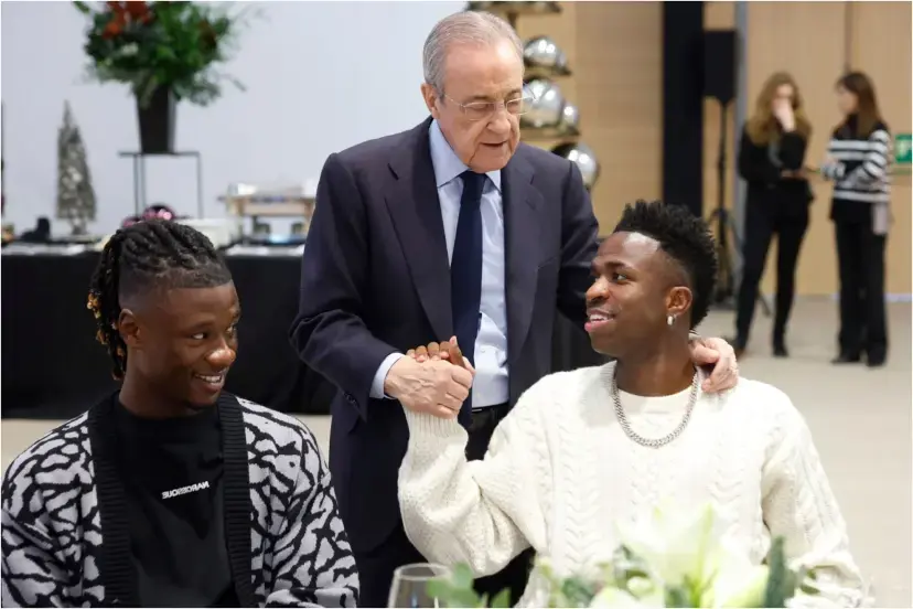 Older club executive stands between two seated footballers during an indoor event, greeting one with a handshake-Vinicius renewal gets Florentino Perez backing as Real Madrid stay calm over future