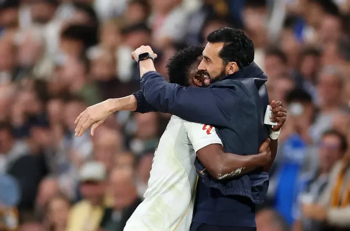 A coach in a dark jacket hugs a Real Madrid player on the pitch after the match